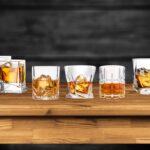 A variety of whiskey glasses—Glencairn, rocks, and lowball—filled with whiskey on a wooden bar counter.